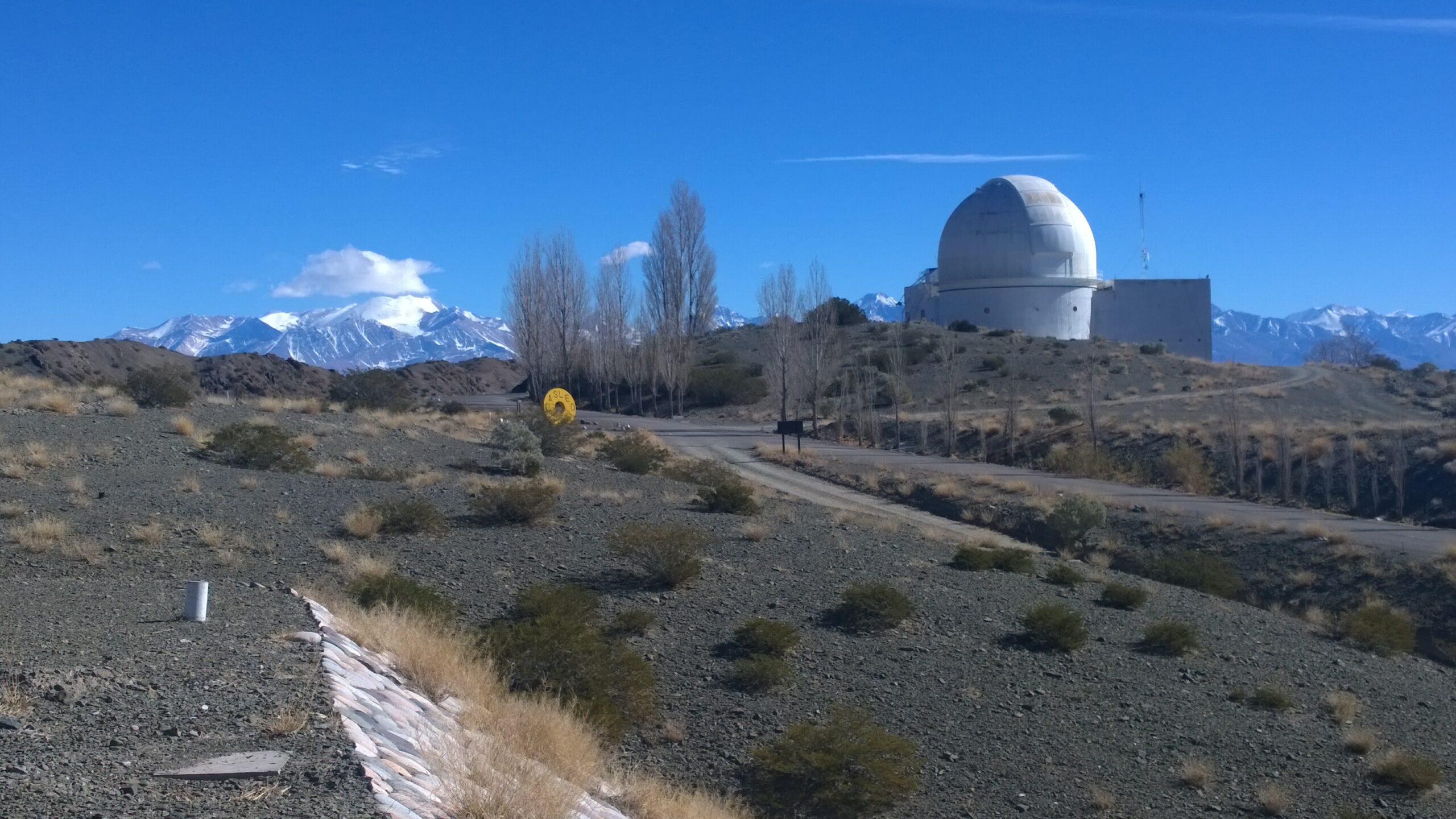 La imagen muestra el Complejo Astronómico El Leoncito. Se observa un paisaje con la Cordillera de los Andes de fondo, un camino y a su derecha, la cúpula del telescopio principal del observatorio.